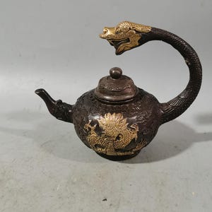 Può includere: Una teiera in bronzo con manico e beccuccio a forma di drago. Il manico è curvo, con la testa del drago all'estremità. Il corpo della teiera è decorato con un disegno di drago dorato.