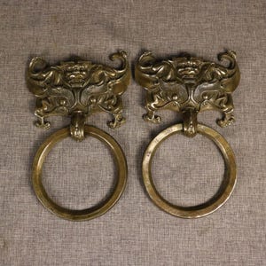 May include: Two antique bronze door knockers. Each knocker features a detailed, ornate design with a central face and stylized wings. The ring handles are attached below. The bronze has a warm, aged patina.