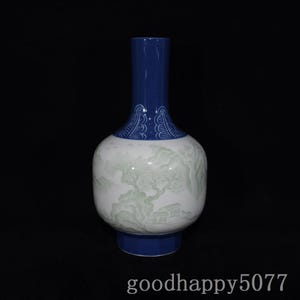 May include: A ceramic vase with a tall, solid blue neck and base. The bulbous body is white and features a pale green landscape illustration depicting trees, mountains, and small structures. The text "goodhappy5077" is printed at the bottom.