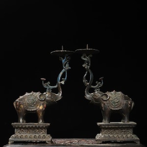 May include: Two bronze elephant candle holders. Each elephant stands on a decorative base and supports a candle cup with a curved, blue-accented stem. The elephants face each other, creating a symmetrical design. The candle holders have an antique finish.