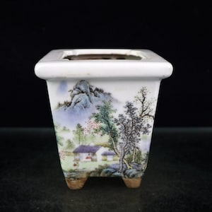 Puede incluir: Una maceta de bonsái de cerámica blanca con forma cuadrada y cuatro pequeñas patas. La maceta está decorada con una escena de paisaje pintada a mano que representa montañas, árboles y una pequeña casa.