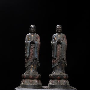Puede incluir: Dos estatuas de bronce de monjes budistas en pose meditativa, con las manos juntas en oración. Las figuras visten túnicas con detalles intrincados y se alzan sobre bases de flores de loto. Las estatuas tienen una pátina oscura con toques de rojo y azul.