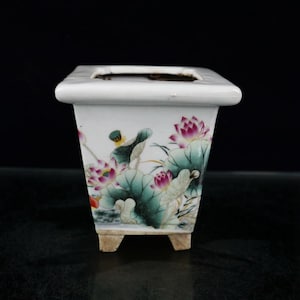 May include: A white ceramic square planter with a floral design featuring pink and green lotus flowers. The planter has four small feet.