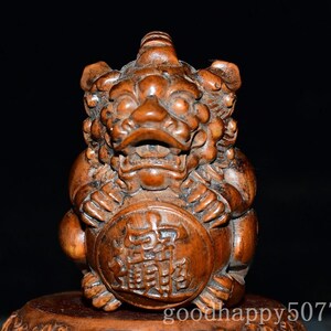 May include: A brown carved wooden figurine of a mythical creature with a wide open mouth and bulging eyes. The creature is holding a circular disc in its front paws, which features intricate carved Chinese characters on its surface.
