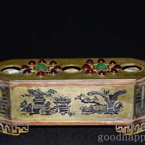 May include: A rectangular brass container with three circular openings on the top, decorated with two floral motifs featuring central green stones surrounded by red stones. The sides display dark blue etched illustrations of vases and plants.