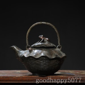 May include: A dark brown metal teapot featuring a textured basket-weave base and a lid shaped like a lotus leaf. A small frog figure sits atop the lid, and a twisted metal handle arches over the top. A small snail is visible near the spout.