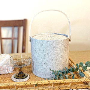 May include: A white and gray speckled ice bucket with a clear plastic handle. The bucket is sitting on a woven wicker tray with a green plant and a clear glass.