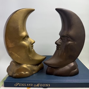 May include: Two crescent moon sculptures with human faces, one gold-toned and the other bronze, sit side-by-side. The sculptures rest on a stack of books, with the titles "Suns and Moons" and "Crystals and healing stones" visible.