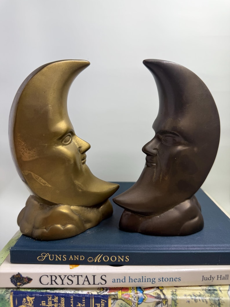 May include: Two crescent moon sculptures with human faces, one gold-toned and the other bronze, sit side-by-side. The sculptures rest on a stack of books, with the titles "Suns and Moons" and "Crystals and healing stones" visible.