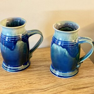 May include: Two blue and green ceramic mugs with white accents. The mugs have a rounded body and a handle.