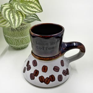 May include: A ceramic coffee mug with a dark brown interior and handle, featuring the text "I take my coffee." The mug has a white base decorated with brown coffee bean designs. A green plant in a decorative pot is in the background.