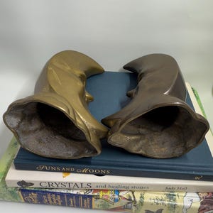 May include: Two brass-toned sculptures, each with a curved, hooded shape and a visible profile. The sculptures rest on a stack of books, including one titled "Funs and Moons" and another "Crystals and healing stones". The background is a plain white.