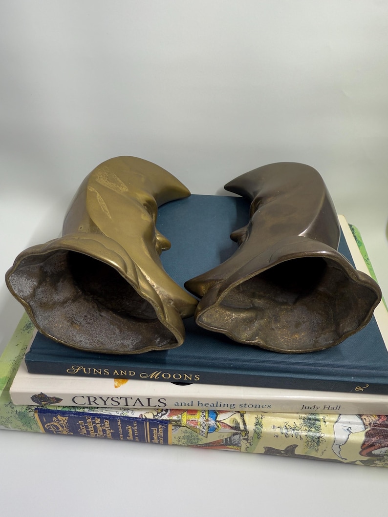 May include: Two brass-toned sculptures, each with a curved, hooded shape and a visible profile. The sculptures rest on a stack of books, including one titled "Funs and Moons" and another "Crystals and healing stones". The background is a plain white.