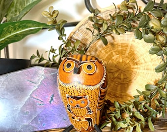May include: A hand-carved wooden owl figurine with intricate details. The owl is brown and orange with black eyes and a small owl carved on its chest. The figurine is standing on a wooden base.