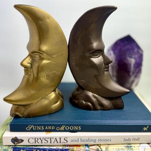 May include: Two crescent moon bookends, one gold and one bronze, with human faces. They rest on a stack of books, including "Suns and Moons" and "Crystals and healing stones." A purple crystal is in the background.