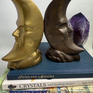 May include: Two crescent moon bookends with human faces, one gold-toned and one bronze-toned, stand on a stack of books. A large purple amethyst crystal is placed between the bookends. The books include titles like "Suns and Moons" and "Crystals and healing stones."