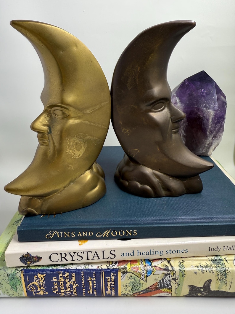 May include: Two crescent moon bookends with human faces, one gold-toned and one bronze-toned, stand on a stack of books. A large purple amethyst crystal is placed between the bookends. The books include titles like "Suns and Moons" and "Crystals and healing stones."