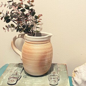 May include: A light brown ceramic pitcher with a white rim and a handle. The pitcher is filled with a sprig of eucalyptus with green leaves and pink stems. The pitcher is sitting on a light blue and green wooden surface.