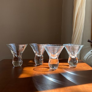 May include: Four clear glass martini glasses with a unique design. Each glass has a flared bowl and a rounded base, sitting on a wooden surface. The glasses are arranged in a row, with sunlight casting shadows.