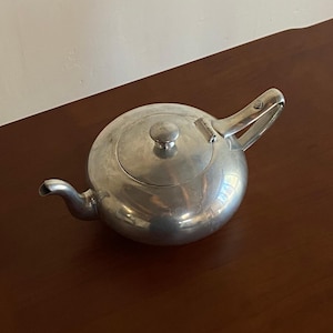 May include: A silver-colored teapot with a rounded body, a curved spout, and a hinged handle. The lid has a small, round knob. The teapot is made of metal and sits on a dark brown wooden surface.
