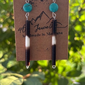 May include: Dangle earrings featuring a round turquoise bead with dark veining above a long, cylindrical quill-like bead that transitions from white at the top to dark brown at the bottom, suspended from silver-toned metal hooks on a brown card.