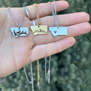 May include: Three Montana state-shaped necklaces with different designs. One is silver with a deer, another is gold with mountains, and the third is silver with a heart. Each necklace hangs from a delicate chain.
