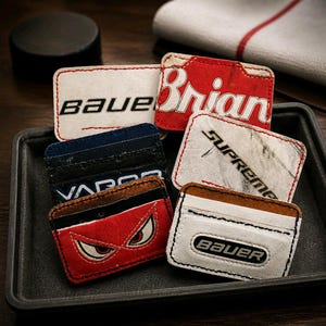 Könnte beinhalten: Eine Sammlung rechteckiger Kartenetuis aus wiederverwerteter Eishockeyausrüstung mit verschiedenen Logos und Grafiken wie Bauer, Brian's, Supreme und Vapor, versehen mit genähten Kanten und markanten Farbschemata in Rot, Weiß und Blau.