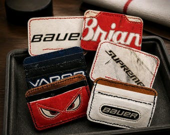 Peut inclure: Une collection de porte-cartes rectangulaires fabriqués à partir d'équipements de hockey recyclés, arborant divers logos et graphismes tels que Bauer, Brian's, Supreme et Vapor, avec des bords cousus et des coloris distincts rouge, blanc et bleu.
