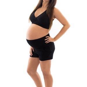 Puede incluir: Una mujer embarazada con un sujetador deportivo negro y pantalones cortos negros. Est&aacute; de pie con la mano en la cadera y sonriendo a la c&aacute;mara.