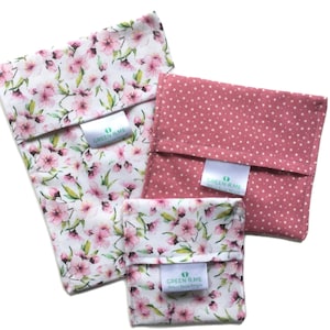 May include: Three reusable food storage bags in different sizes and patterns. The largest bag is white with a pink floral pattern, the medium bag is pink with white stars, and the smallest bag is white with a pink floral pattern. All bags have a green label with the text "GREEN-R-ME Reusable Food Bags".