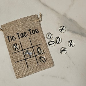 May include: A burlap bag with the words "Tic Tac Toe" printed on it. The bag contains nine glass game pieces with either an "X" or an "O" printed on them.