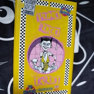 Könnte beinhalten: Eine gelbe Tarotkartenbox mit den Worten "Pink Rock Tarot" in rosa Schrift. Die Box zeigt eine Cartoon-Illustration einer Person mit Irokesenschnitt und dem Text "Never Mind Solos". Die Box hat auch einen Warnhinweis für Eltern.