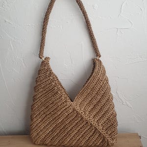 May include: A tan crochet handbag featuring a diagonal ribbed texture and a single rounded handle attached with gold-toned metal rings. The bag has a triangular silhouette with a V-shaped opening at the top, constructed from a thick, durable yarn.