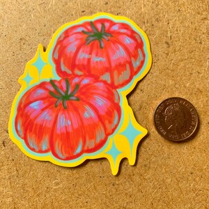 K&ouml;nnte beinhalten: Ein Aufkleber mit zwei roten Tomaten mit gr&uuml;nen Stielen und blauen Highlights, umrandet in Gelb mit blauen Sternen-Akzenten. Eine M&uuml;nze befindet sich neben dem Aufkleber.
