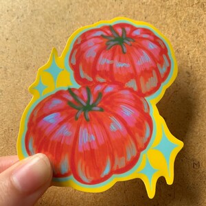 K&ouml;nnte beinhalten: Ein Aufkleber mit zwei roten Tomaten mit gr&uuml;nen Stielen, umrandet in Blau und Grau, vor einem gelben Hintergrund mit blauen Sternen-Akzenten. Der Aufkleber wird von einer Hand gehalten.