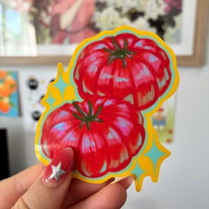 K&ouml;nnte beinhalten: Ein Aufkleber mit zwei roten Tomaten mit gr&uuml;nen Stielen, umrandet in Gelb mit t&uuml;rkisfarbenen Sternen. Die Tomaten haben rosa und blaue Highlights, was ihnen ein lebendiges, illustriertes Aussehen verleiht. Der Aufkleber wird von einer Hand mit einer sternf&ouml;rmigen Nageldekoration gehalten.