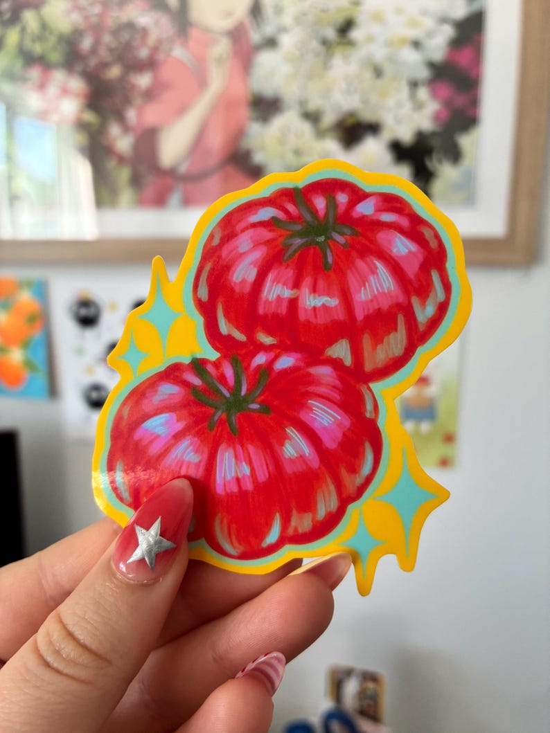 K&ouml;nnte beinhalten: Ein Aufkleber mit zwei roten Tomaten mit gr&uuml;nen Stielen, umrandet in Gelb mit t&uuml;rkisfarbenen Sternen. Die Tomaten haben rosa und blaue Highlights, was ihnen ein lebendiges, illustriertes Aussehen verleiht. Der Aufkleber wird von einer Hand mit einer sternf&ouml;rmigen Nageldekoration gehalten.