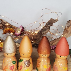 Könnte beinhalten: Vier Holzpilzfiguren mit bemalten Blatt- und Punktdetails. Jede hat eine unterschiedlich farbige Kappe: Silber, Orange, Braun und Rot. Die Figuren sind auf einer Holzoberfläche angeordnet, mit getrockneten Blumen und Lichterketten im Hintergrund.