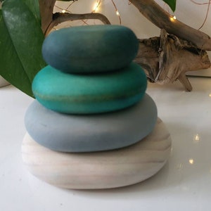 Puede incluir: Una pila de cuatro piedras de madera pintadas en tonos azules y verdes. La piedra inferior es de color beige claro, seguida de una gris claro, una turquesa y una piedra verde oscuro en la parte superior. Las piedras están apiladas una encima de otra.