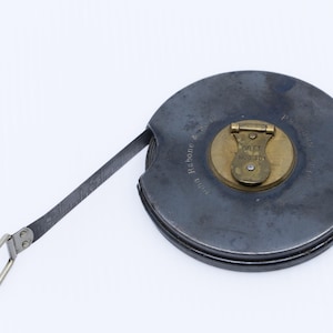 Puede incluir: Una cinta métrica de metal vintage con una carcasa gris oscuro y un centro de latón. La cinta se extiende desde la carcasa, con un lazo de metal en el extremo. La carcasa tiene texto alrededor del borde.