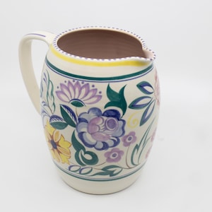 Puede incluir: Jarra de cerámica color crema con un asa grande y un diseño floral. La jarra presenta un borde con una franja amarilla y una franja verde oscuro. El diseño floral incluye flores moradas, azules, amarillas y rosas con hojas verdes.