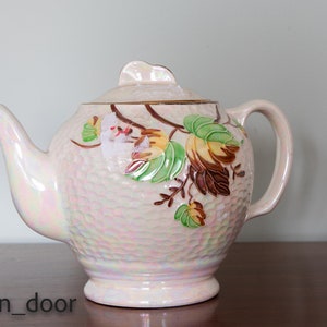May include: A white teapot with a floral design. The teapot has a pearlescent finish and a textured surface. The flowers are in shades of pink, green, and brown.
