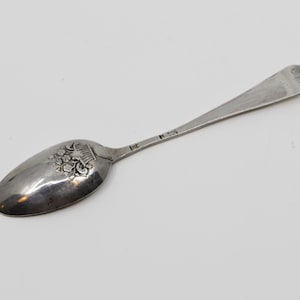 Könnte beinhalten: Ein silberner Löffel mit floralem Design auf der Laffe. Der Löffel hat einen langen Stiel und eine kleine Inschrift am Ende. Der Löffel ist wahrscheinlich Vintage oder antik.
