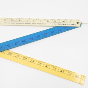 May include: A folding ruler set consisting of three connected segments in white, blue, and yellow. The white segment features black markings from 1 to 12 inches and text reading For the finest selection of patterns, fabrics and sewing accessories, shop WOOLWORTH / Woolco.