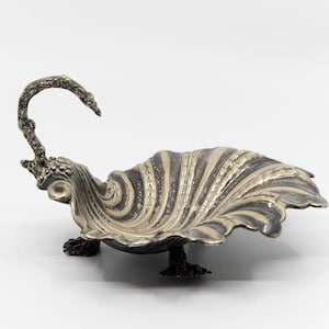 May include: A silver-toned, leaf-shaped dish with a curved handle. The dish has a textured, ridged surface and rests on three small feet. The handle curves upwards, adding a decorative element to the piece.