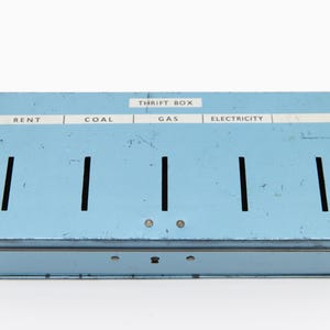 Op de afbeelding: Een vintage lichtblauwe metalen spaarpot met sleuven voor spaargeld. Op de doos staan de woorden "RENT", "COAL", "GAS" en "ELECTRICITY". De doos is rechthoekig en heeft een scharnierend deksel.