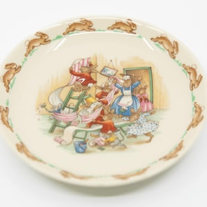 May include: A cream-colored ceramic plate with a whimsical illustration of rabbits. The plate's border features a repeating pattern of brown rabbits. The central image depicts rabbits in a domestic scene, with a ladder and various objects.