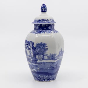 May include: A white and blue ceramic vase with a lid. The vase features a detailed landscape scene in blue, with trees, buildings, and figures. The vase is approximately 12 inches tall.