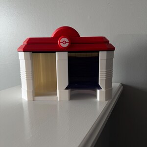 May include: A 3D-printed model of a building, featuring a red roof with a white and red logo, and white walls. The building has two sections, one with a clear window and the other with a dark blue interior. The model is sitting on a white surface.
