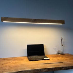Może przedstawiać: Drewniana, prostokątna lampa wisząca z jasnym, liniowym źr&oacute;dłem światła LED. Światło wisi nad drewnianym biurkiem z laptopem i elementami dekoracyjnymi. Biurko wykonane jest z jasnego drewna.